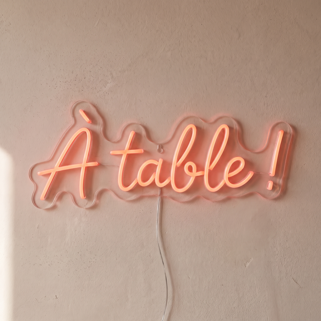 Gros plan du néon mural « À table ! » sur fond beige, tube allumé