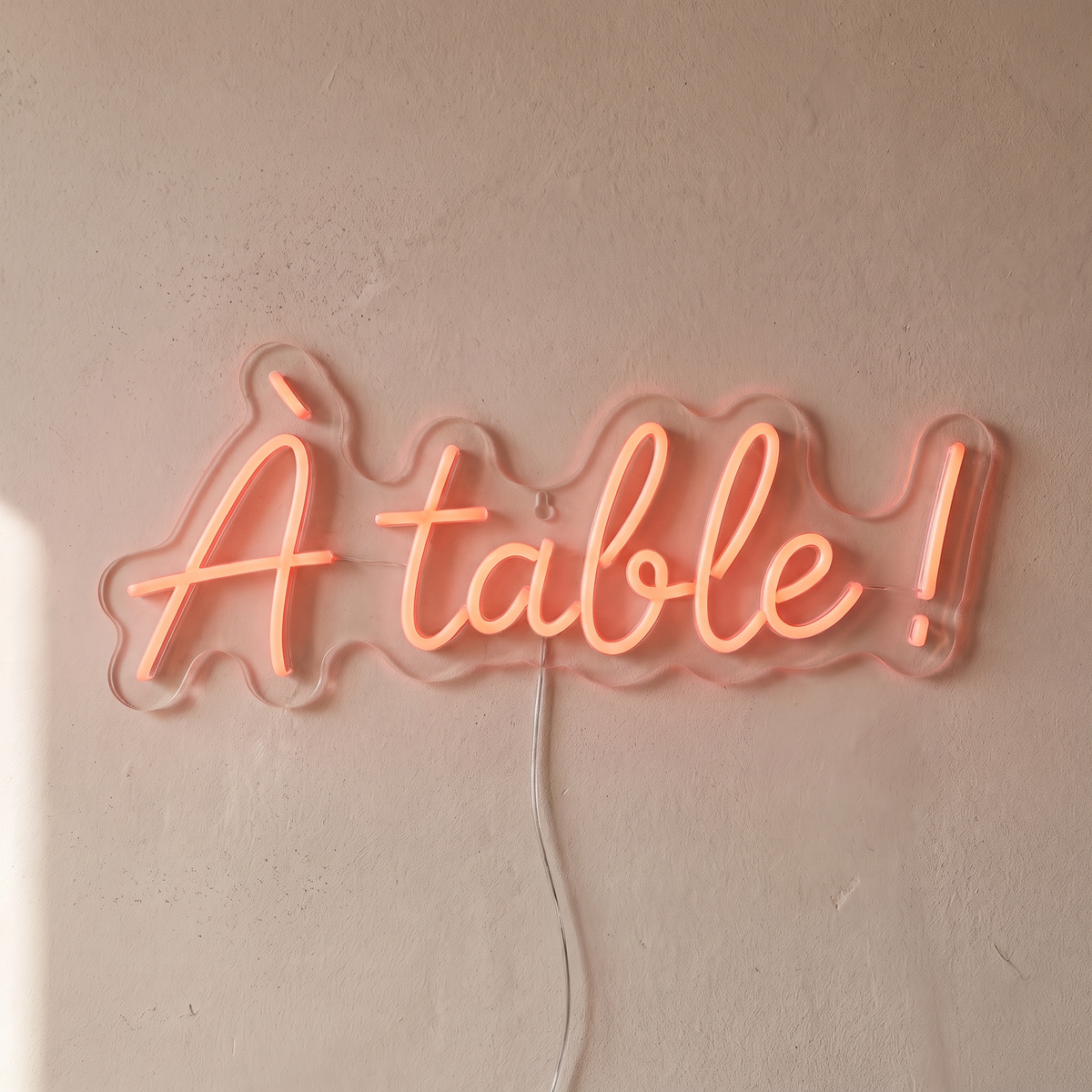 Gros plan du néon mural « À table ! » sur fond beige, tube allumé