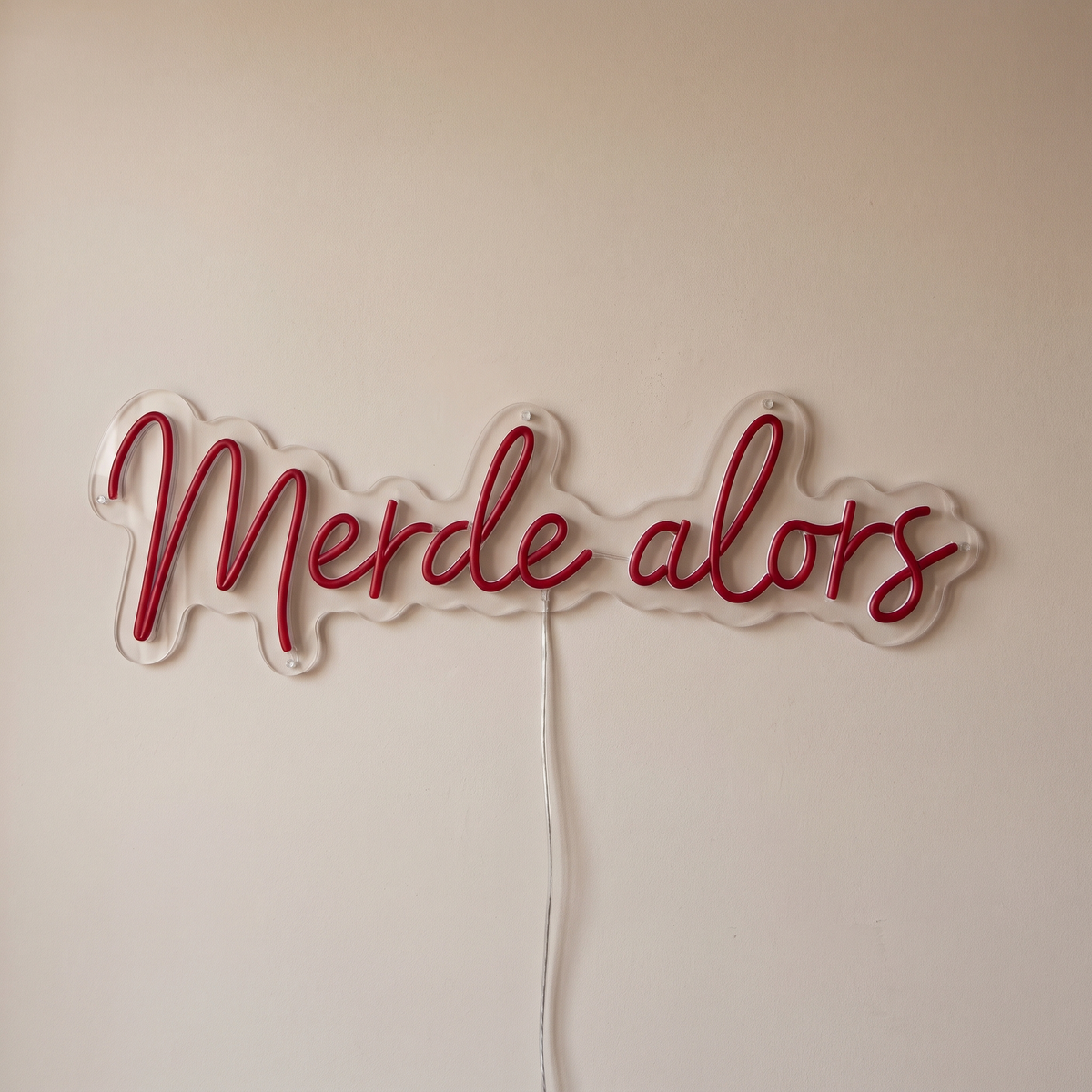 Gros plan du néon mural « Merde alors » sur fond beige, tube allumé